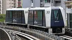 A two-car Mitsubishi Crystal Mover train on elevated tracks. The train is white and dark blue with "SBS Transit" written on the side, and residential buildings are visible in the background.