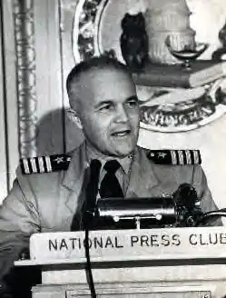 Middle-aged man wearing U.S. Navy khaki uniform and Captain shoulder epaulettes standing behind a lectern with microphone and placard that reads "National Press Club".
