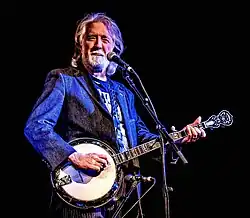 John McEuen, singing into a microphone while playing a banjo