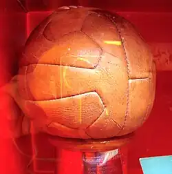 12-panel brown leather football on a plinth of hardwood, with a brass panel inscribed with the names of the victorious team members, photographed through the glass of a display case