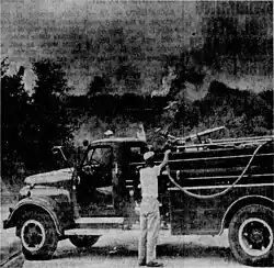 Grainy, black and white newspaper photograph of a man in light clothing operating a hose mounted on a truck as the hillside behind him burns