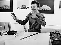 This photograph is of the artist Laurie Anderson with her arms up speaking inside a print shop with artwork hanging on the wall behind her.