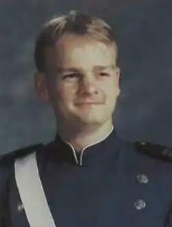 A color portrait photo of a white young man, he is wearing a blue academy uniform, smiling while facing and looking slightly to the camera's right