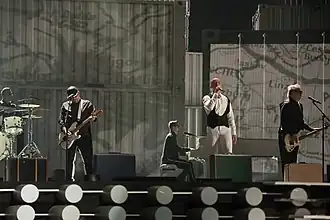 Several men in suits performing on a stage in front of a grey backdrop.