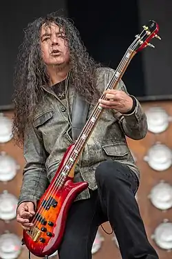 A man with long black hair wearing a leather jacket and playing a bass guitar.