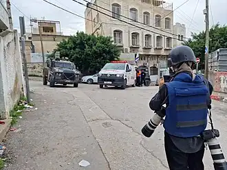 Israeli soldier standing in the street near an intersection with a hospital building in front of the soldier and a military vehicle and ambulance in the intersection.