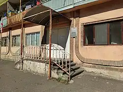 The entrance to an old, two-story, peach-colored building. A rusty metal awning covers the entrance, which is reached by concrete steps with peeling white railings. A sign is mounted on the wall to the right of the door.