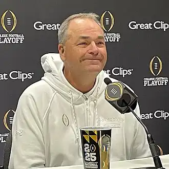 A football coach smiles in front of a microphone.
