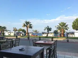 This photograph depicts a portion of the annual event held in Nea Marki, where books and pieces of folk art are sold in booths under tents. The photograph is from inside a restaurant across the street from the fair. The beach is visible behind the fair. The photograph was taken in 2023.
