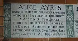A tablet formed of six standard sized tiles, bordered by green flowers in the style of the Arts and Crafts movement. The tablet reads "Alice Ayres, daughter of a bricklayer's labourer who by intrepid conduct saved 3 children from a burning house in Union Street, Borough, at the cost of her own young life April 24, 1885".
