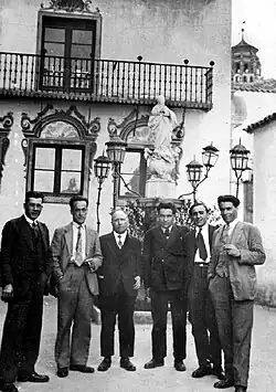 Men in suits standing in front of a statute and a traditional-style Spanish building