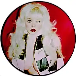A circular picture disc of Angelyne wearing a checkerboard jacket against a red background