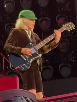 Angus, aged 69, is shown wearing his trademark schoolboy uniform. He faces away to his left, plays his Gibson SG guitar.