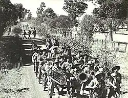 Soldiers marching along a dirt road in the Australian bush, led by a brass band