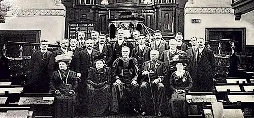 A black and white photo of about 20 people inside a church. The front row are seated, while two additional rows of people behind them are standing.