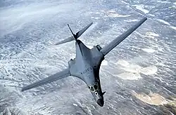 Top forward view of gray aircraft with wings swept forward banking right. Underneath are strips of white clouds and uninhabited terrain.