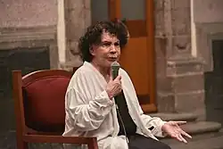 A sitting woman wearing black and white clothes speaking at a microphone in public.