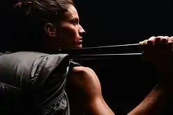 A woman stands in shadows, wearing a hiking backpack and gripping two hiking sticks slung over her shoulder. She looks off to the side, while warm light highlights her muscles and face.