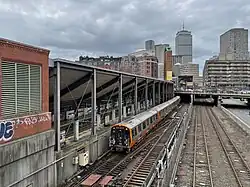 A railway cut in a city. At left are three tracks with platforms under a large trainshed. At center is a two-track line with a metro track. At right is a two-track line.