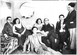 A black and white photograph of 7 people in a room. From left to right, a white man with glasses lying on a sofa; a white woman lying on the man and looking away; a white grinning man in a suit sitting on the floor; the upper body of a woman passing her arm over the shoulders of the man on the right; an older white man sitting on the sofa in a suit; a white man with dark made-up lips sitting on a chair; a standing white man laying his hand on the sitting man's shoulder.