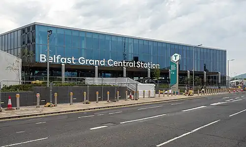 An image of a large train station with large glass windows and a Translink logo atop a green and grey sign beside a set of stairs