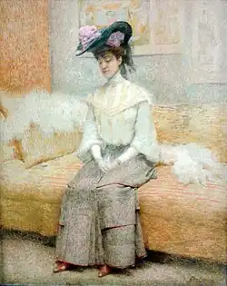 A seated woman rests on a sofa, looking downwards with a contemplative or demure expression. She wears a light-colored top with long sleeves and a high collar, a flowing grey or light brown skirt, and a floral hat with ribbons. A feathery shawl drapes around her shoulders and across the sofa. The background suggests a softly colored interior with patterned wallpaper or textiles.