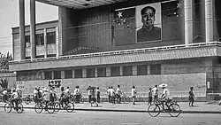 A black and white photo of a street in China in the 1970s. Cyclists bike past a large building with Big Character Posters and a portrait of Mao Zedong