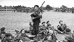 Black-and-white photograph of Millin in uniform, standing and playing his bagpipe, while uniformed soldiers lay and lounge around him on the grass in a field