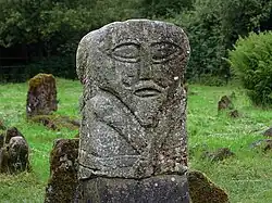 Photograph of the figure in-situ in Boa island's Caldragh graveyard. The oval face has big eyes, a straight nose and a half-open mouth. Its body extends from just above the waist.