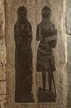 This image shows a medieval monumental brass depicting Sir John and Lady de Creke, engraved into a large stone slab set into a church floor. The brass shows two full-length figures standing side by side: on the left, Lady de Creke is portrayed in typical 14th-century female dress with a flowing gown, veil, and wimple, her hands positioned in prayer. On the right, Sir John de Creke is shown in full military attire including a mail hauberk and coif, with his hands also clasped in prayer. The knight's detailed armour includes a great helm and sword, whilst decorative elements and heraldic devices are visible at the base of the figures. The brass demonstrates the fine engraving technique typical of English monumental brasses, with intricate linear detail clearly visible against the dark metal plates set into the pale limestone matrix. This type of memorial became popular from the 13th century onwards as a more affordable alternative to sculptural effigies.