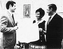 Three people holding a large newspaper or document standing in front of a small framed piece of art.