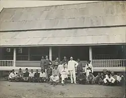Sepia photograph of a number of people in front of a building with a corrugated iron roof