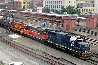 A freight train passing through a train station, lead by three diesel locomotives in various paint schemes.