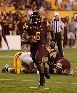 An American football player in a dark red and yellow uniform cradles the football as he sprints towards the camera. Two players in the background lie on the ground.