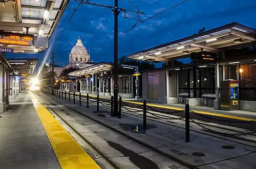 A low-angle shot of a side-platform two-track at-grade train station in the evening. In the background, an illuminated Italian Renaissance-style dome.