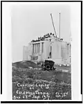 The Cardston temple at a distance, as if on a postcard. People can be seen at a distance standing on top of the structure, an old car sits in the middle of the shot, sitting on a hill, among other hills of dirt.