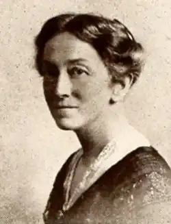 A middle-aged white woman with darkish hair dreseed to the nape, wearing a dark dress