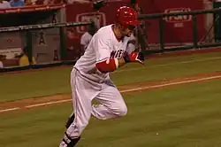 A dark-skinned man with "NGEL" visible in red text on a white jersey runs from home plate to first base. He has on red and black batting gloves, a red batting helmet, white pants and black shinguards.