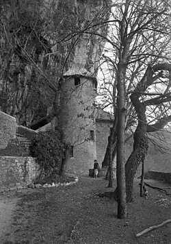 Early view of a castle tower against a rock face, with a courtyard planted with some trees in front.