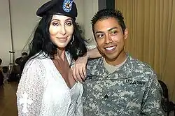 Cher wears a military beret and a white lace outfit, smiling with her arm around a US Army soldier in uniform.