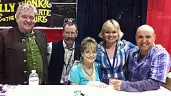 Five middle-aged white adults (two women and three men) in casual dress are behind a table, posing for a group photo.