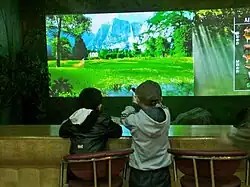 Two children, one sitting down and another one standing while aiming, are playing a light-gun shooter game in front of a large projected screen. It displays a forest area and appears to feature several animals which one must shoot to achieve points.