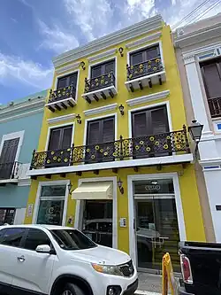 Photograph of the exterior of a building; there is a yellow building with 3 stories and a balcony on the second level.