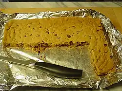 A foil-lined tray with a solid light brown sheet with dark brown spots. A section has been cut away and most of a knife is lying in that portion.