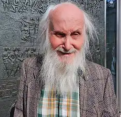 Colour Press headshot of a smiling, elderly man with long white hair and beard, wearing a tweed suit and plaid scarf