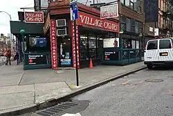 View of the triangle, which is located on a sidewalk at a street corner. The triangle is outside the Village Cigars shop and the Christopher Street–Sheridan Square station of the New York City Subway. The triangle can be seen on the sidewalk toward the left side of the photo.