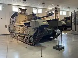 Three-quarter right-front view of a tracked armoured vehicle with gun barrel and turret in situ inside a large museum gallery.