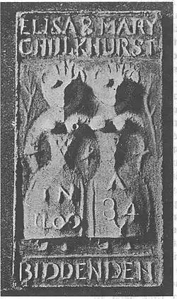 Two highly simplified female figures standing side by side, with snout-like faces, conical exposed breasts, extremely narrow waists and very wide hips. The figures each only have one arm, and appear to be joined at the shoulder. Crown-like points rise from their heads. Next to each figure is a branch. The words "Eliza & Mary Chulkhurst" are written above the figures, and the word "Biddenden" is written below them. The left figure has "In 1100" written on her dress, and the right figure has "A 34" written on her dress.