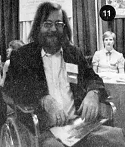 Grainy black and white photograph of a bearded man in glasses sitting in a wheelchair.