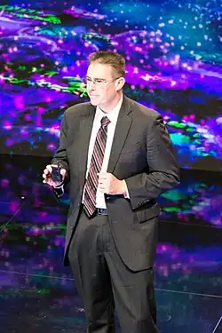 Rob Knight wearing a light-coloured shirt, striped tie, and grey suit, standing onstage, holding a presentation remote controller in front of a shimmery, multicoloured background, appearing to speak into a headset microphone